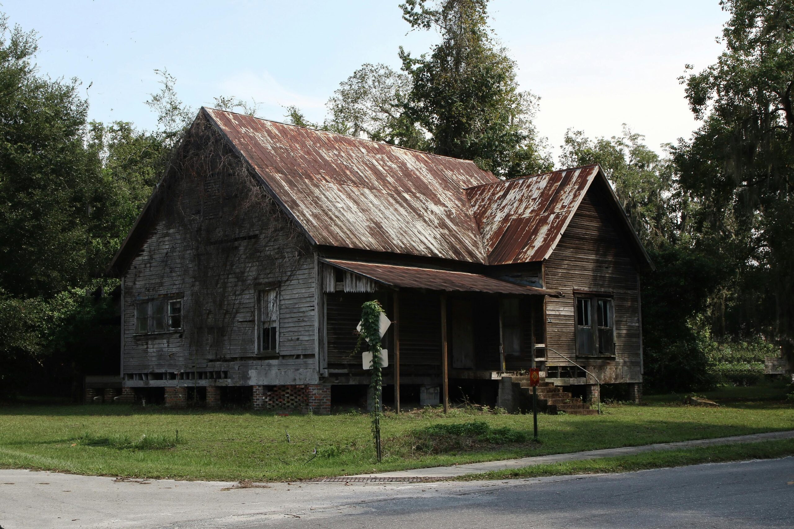 An old, weathered house with a rusty roof stands in an overgrown area surrounded by trees.