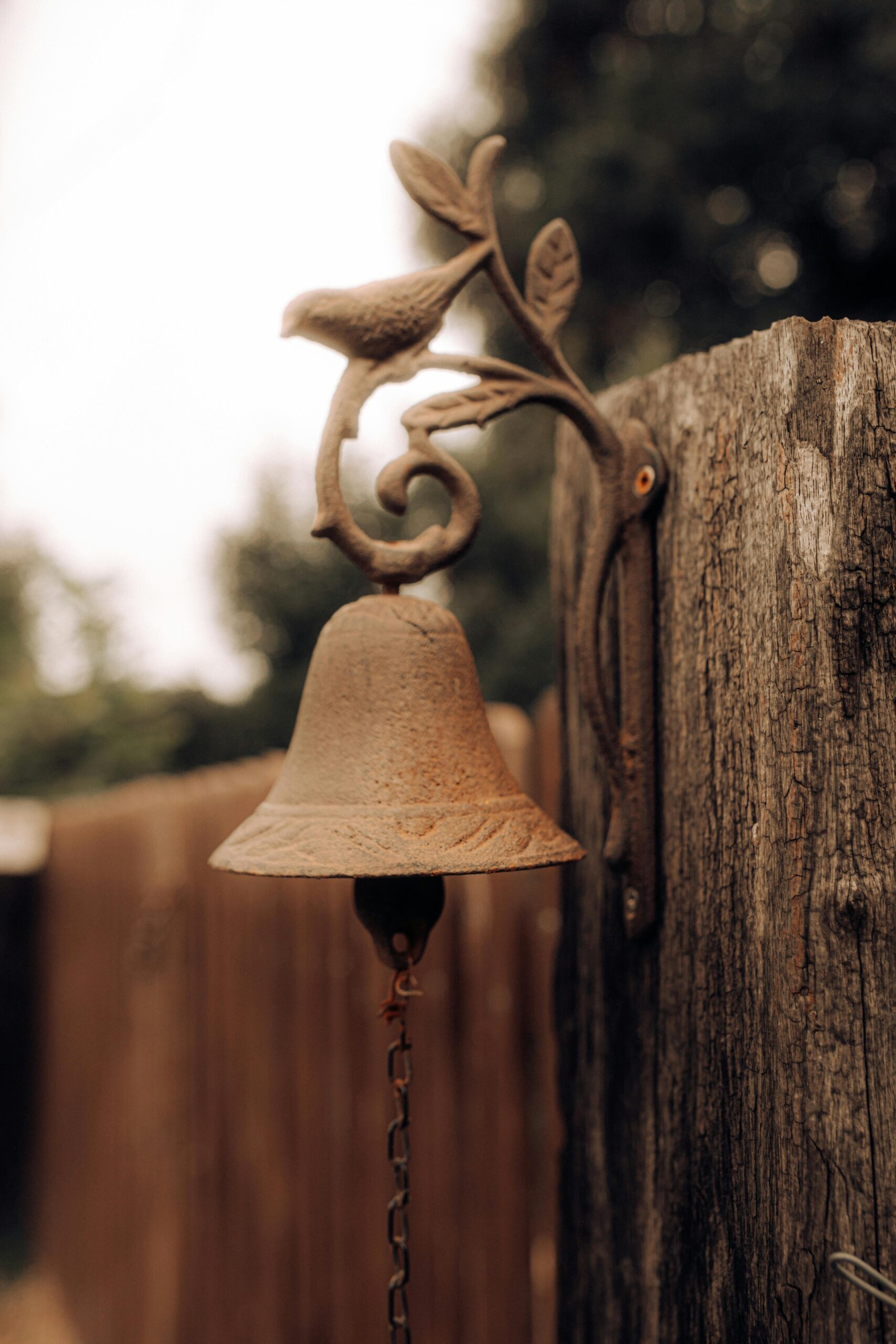 A vintage-style bell with a bird detail, mounted on a rustic wooden fence outdoors.