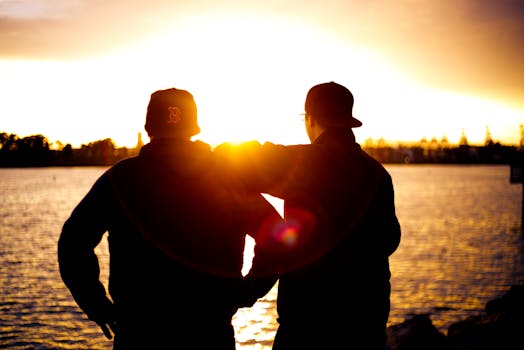 Two friends in baseball caps watch a sunset over the water, creating a serene silhouette.